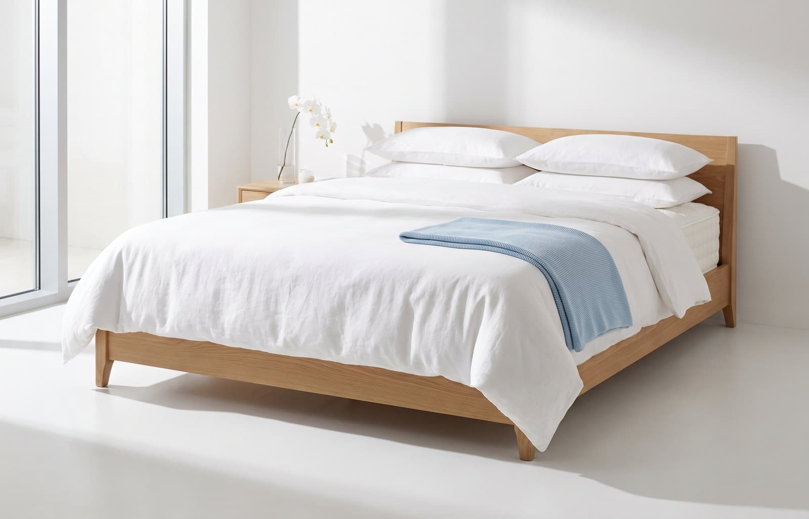 A neatly made bed with white linen and a light oak frame in a sunlit showroom bedroom